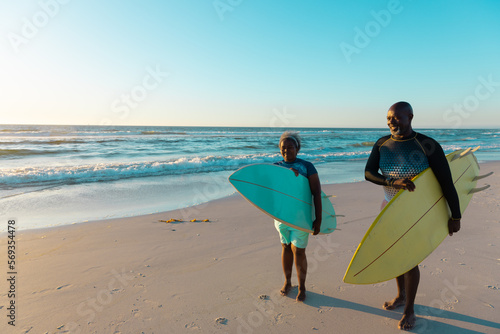 African american senior couple carrying surfboards while walking at sandy beach against sky
