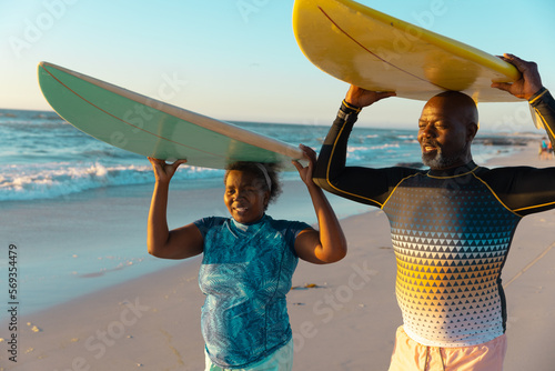 African american senior couple carrying surfboards on heads while standing at beach against sky