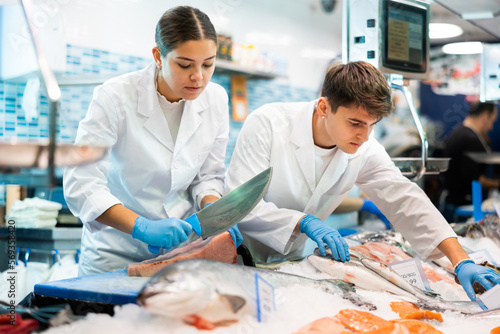 Skilled shop assistants cutting piece of tuna in fish shop with big assortment