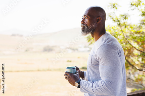 Bald african american senior man with mug contemplating and standing in balcony against clear sky