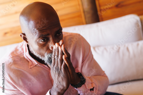 Close-up of worried bald african american senior man with hands clasped sitting on sofa in log cabin photo