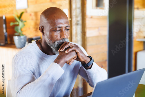 Serious bald african american senior man with hands clasped looking at laptop in log cabin