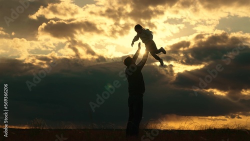 Dad plays with his daughter  throws child up into sky with his hands  happy kid. Father of daughters play together in park against backdrop of sun and clouds. Silhouette of happy family outdoors.