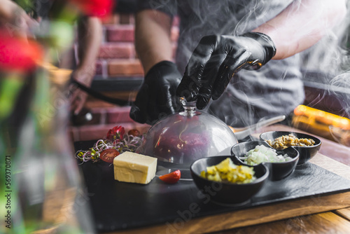 Fototapeta Naklejka Na Ścianę i Meble -  Person in black gloves smoking beef tartare under transparent lid served on black wooden board with garnishes on the side. Polish cuisine. Horizontal indoor shot. High quality photo