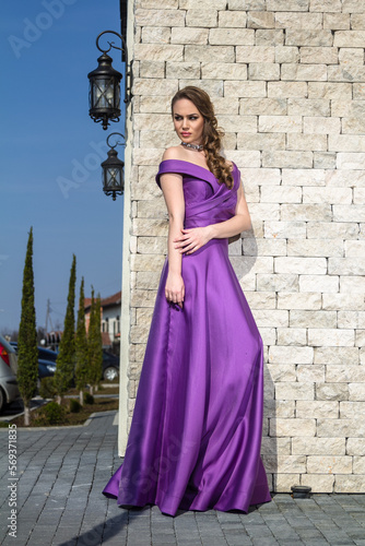 Young woman in a purple dress stands leaning against the wall