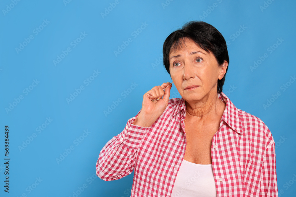 Senior woman cleaning ear with cotton swab on light blue background. Space for text