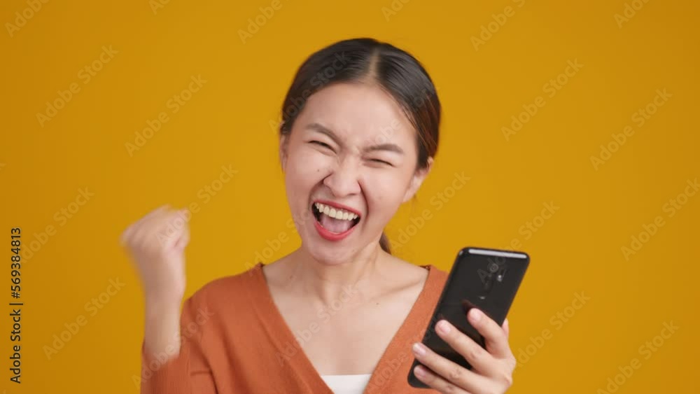 Young Asian woman receive good news from message in smartphone. Excited happy lady scream and very glad on yellow studio background. Young Asian woman receive good news from message in smartphone. Excited happy lady scream and very glad on yellow studio background.