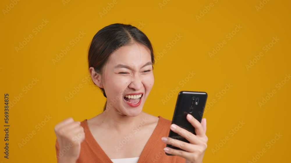 Young Asian woman receive good news from message in smartphone. Excited happy lady scream and very glad on yellow studio background. Young Asian woman receive good news from message in smartphone. Excited happy lady scream and very glad on yellow studio background.