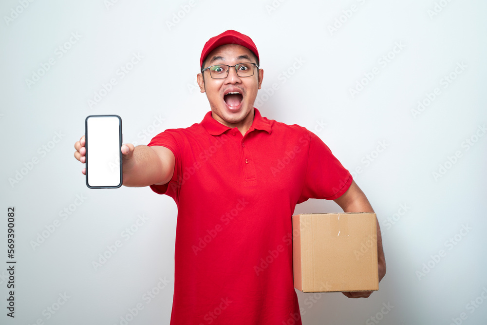Happy young asian delivery man showing blank phone screen in red uniform holding cardboard box Happy young asian delivery man showing blank phone screen in red uniform holding cardboard box