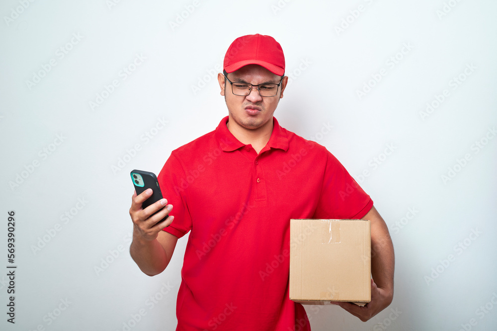 Angry asian courier man in red uniform look at his smartphone while holding cardboard box parcel isolated Angry asian courier man in red uniform look at his smartphone while holding cardboard box parcel isolated
