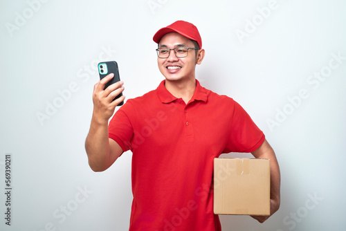Asian courier man in red uniform smiling look at his smartphone while holding cardboard box parcel