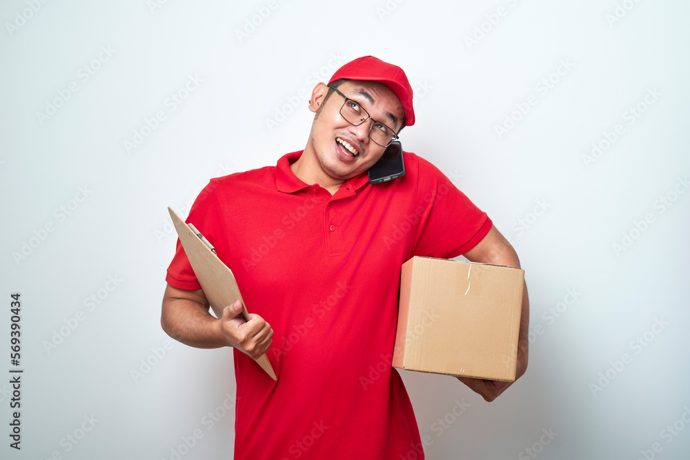 Smiling busy asian courier man answering phone call, holding mobile on shoulder, prepare package for delivery to client and making notes Smiling busy asian courier man answering phone call, holding mobile on shoulder, prepare package for delivery to client and making notes