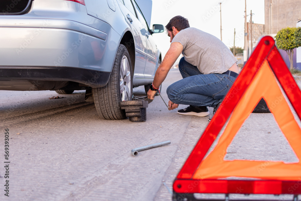 young man changing a flat tire on the road, using a hazard triangle on the road having an emergency young man changing a flat tire on the road, using a hazard triangle on the road having an emergency