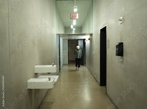 Eerie photo of a generic concrete bathroom hallway with drinking fountains and older woman walking out the door.