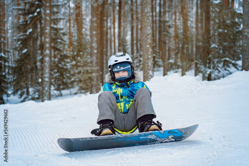 little boy sitting on snow putting his feet in snowboard bindings adjusting straps.