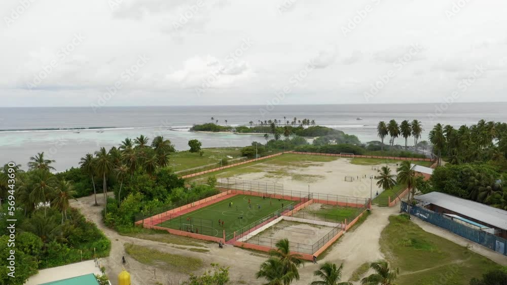 Aerial Panning Shot Of People Playing Soccer On Field Near Sea, Drone Flying Over Landscape Against Cloudy Sky - Thulusdhoo, Maldives Aerial Panning Shot Of People Playing Soccer On Field Near Sea, Drone Flying Over Landscape Against Cloudy Sky - Thulusdhoo, Maldives