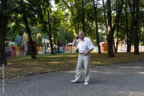An elderly man walks alone in the park in the summer. A modern retired businessman in a white shirt and trousers is talking on a mobile phone © ALA