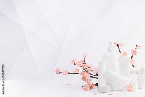 Beauty products for makeup, skin care - cream, lotion in white bottles, branch of spring pink sakura flowers, toiletry in modern light white interior in minimalist geometric japan style, copy space.