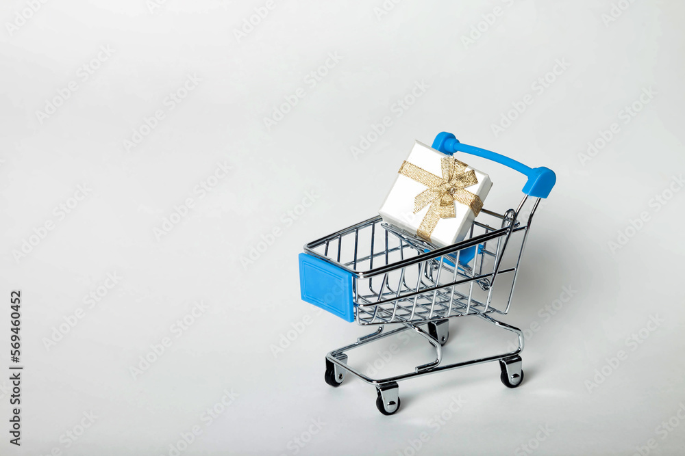 Small empty metal shopping cart with blue handles with gift box present on grey background. Shop buying holiday concept. Copy text space for advertising
