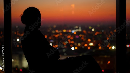 Sad man sits in dark room, holds toy house in his hand and throws it away, turns his head and looks out window at night cityscape. The relevance and problem of housing in context of rising real estate photo