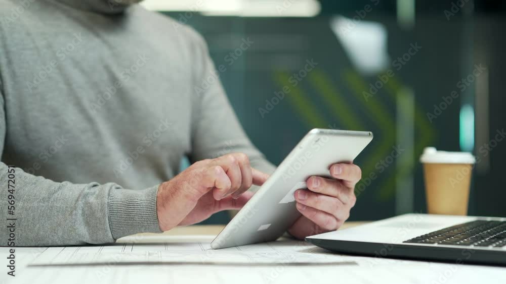 Close up. Man holding a tablet while sitting at a desk at a workplace in a modern office. Employee at work uses the device by moving his finger on the screen, looking for information on the Internet Close up. Man holding a tablet while sitting at a desk at a workplace in a modern office. Employee at work uses the device by moving his finger on the screen, looking for information on the Internet