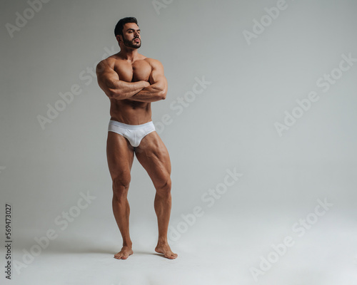 Sexy muscular man in white underwear posing in studio. Handsome bodybuilder standing on grey background. Big male model showing his muscles.