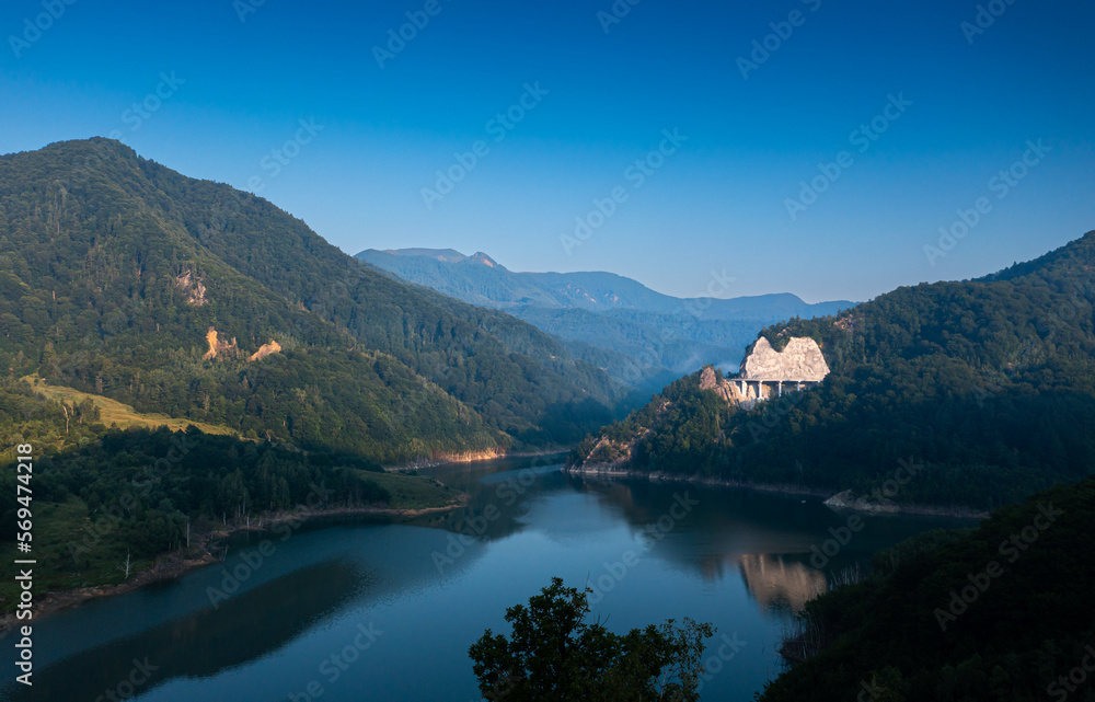 Aerial view of a foggy morning sunrise over a lake and a dam in the mountains next to Siriu Romania, with a cliff road winding