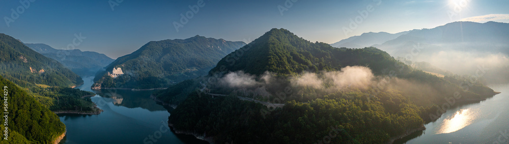 Aerial view of a foggy morning sunrise over a lake and a dam in the mountains next to Siriu Romania, with a cliff road winding