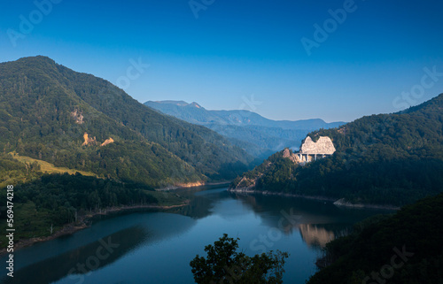 Aerial view of a foggy morning sunrise over a lake and a dam in the mountains next to Siriu Romania, with a cliff road winding © Dragoș Asaftei