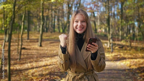 Front view of attractive, young lady standing in park, using smartphone. Glad, happy female reading, texting, wondering, looking at camera. Concept of modern, urban lifestyle.