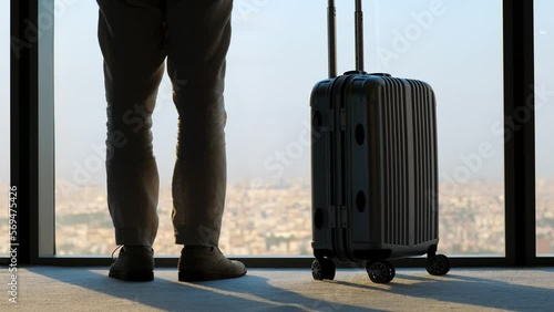 Tourist is nervous whether his flight has been cancelled. Man raise one leg and scratches his calf with foot. Packed suitcase stands next to him. Blurred cityscape seen in background, low half shot