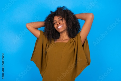 young woman with afro hairstyle wearing brown dress over blue wall stretching arms, relaxed position.