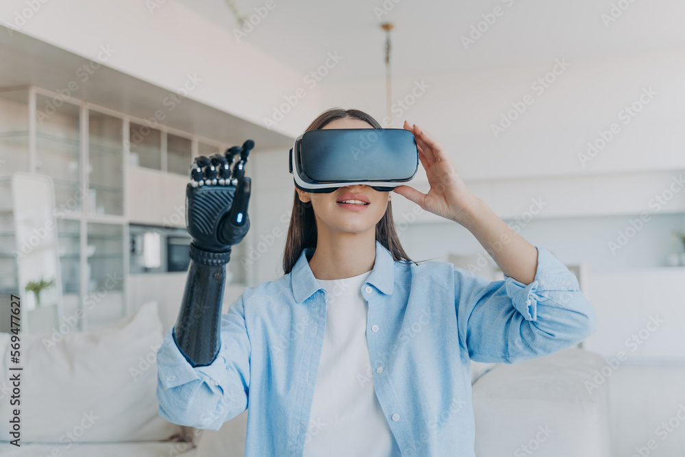 Naklejka premium Disabled person gets rehabilitation. Handicapped girl with cyber arm in vr goggles at home.
