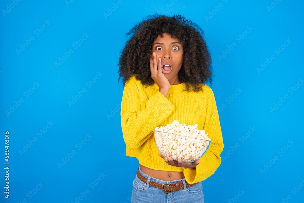 Stupefied young woman with afro hairstyle wearing yellow sweater over blue wall expresses excitement and thrill, keeps jaw dropped, hands on cheeks, has eyes popped out Stupefied young woman with afro hairstyle wearing yellow sweater over blue wall expresses excitement and thrill, keeps jaw dropped, hands on cheeks, has eyes popped out