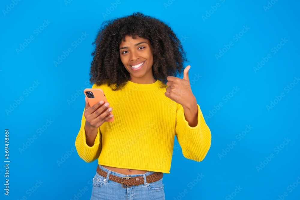 Portrait of young woman with afro hairstyle wearing orange crop top over blue wall using and texting with smartphone happy with big smile doing ok sign, thumb up with fingers, excellent sign Portrait of young woman with afro hairstyle wearing orange crop top over blue wall using and texting with smartphone happy with big smile doing ok sign, thumb up with fingers, excellent sign