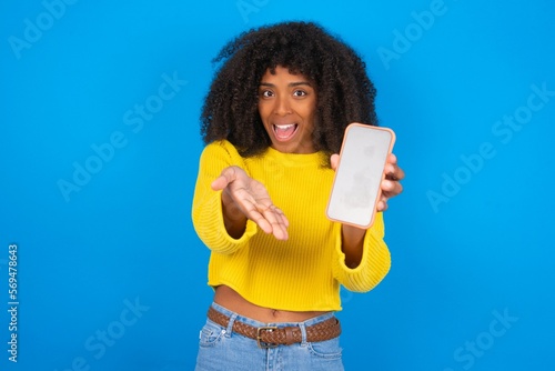 young woman with afro hairstyle wearing orange crop top over blue wall with a mobile. presenting smartphone. Advertisement concept.