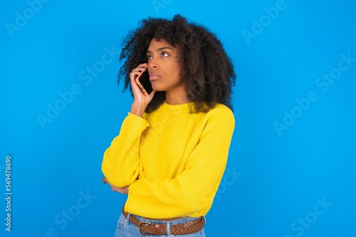 Sad young woman with afro hairstyle wearing orange crop top over blue wall talking on smartphone. Communication concept.