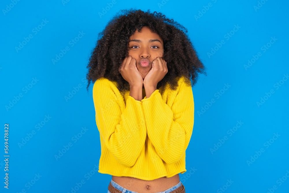 young woman with afro hairstyle wearing orange crop top over blue wall with surprised expression keeps hands under chin keeps lips folded makes funny grimace young woman with afro hairstyle wearing orange crop top over blue wall with surprised expression keeps hands under chin keeps lips folded makes funny grimace