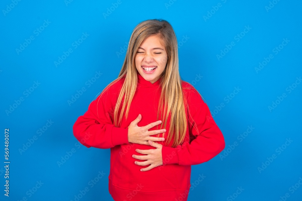caucasian teen girl wearing red sweater over blue studio background keeps hands crossed, laughs out loud at good joke, wears casual clothes. caucasian teen girl wearing red sweater over blue studio background keeps hands crossed, laughs out loud at good joke, wears casual clothes.