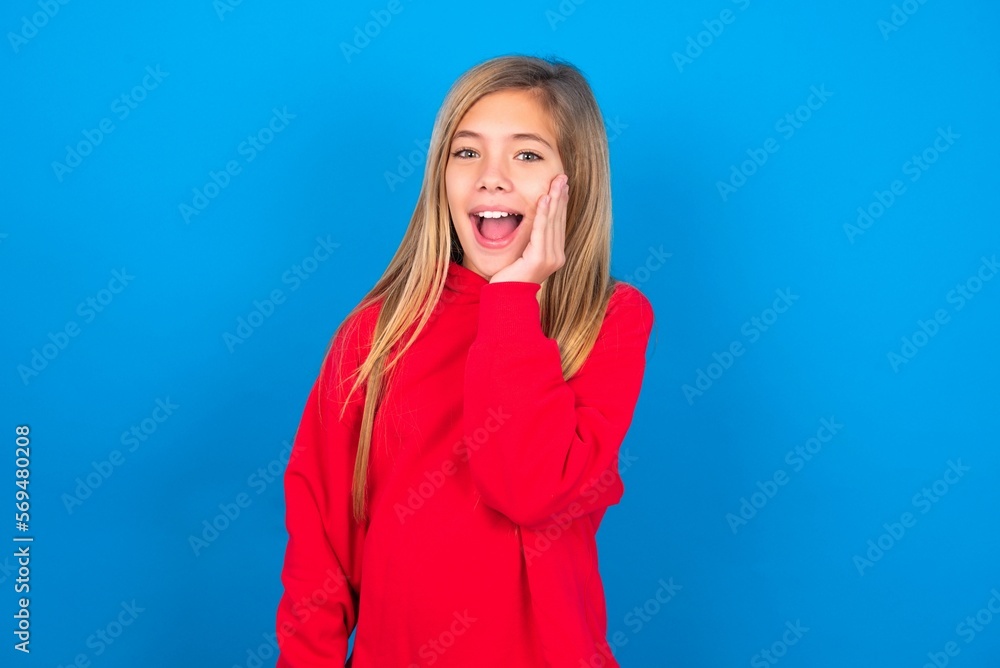 Shocked, astonished caucasian teen girl wearing red sweater over blue wall looking surprised in full disbelief wide open mouth with hand near face. Positive emotion facial expression body language. Shocked, astonished caucasian teen girl wearing red sweater over blue wall looking surprised in full disbelief wide open mouth with hand near face. Positive emotion facial expression body language.