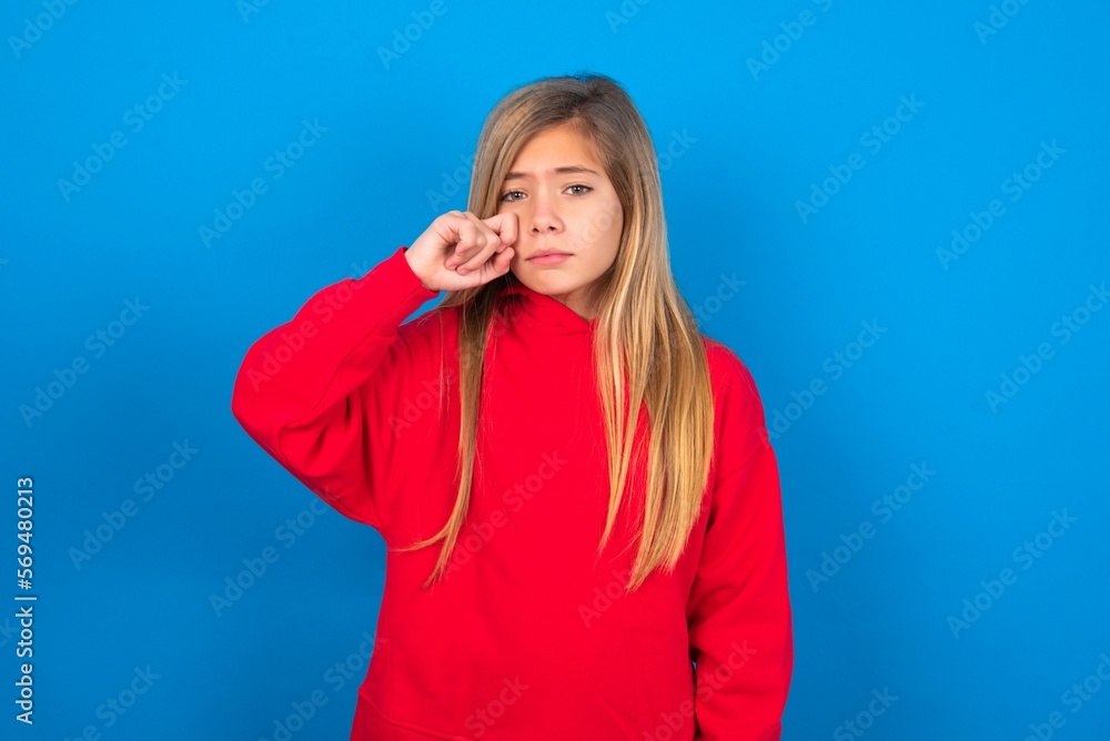 Unhappy caucasian teen girl wearing red sweater over blue studio background crying while posing at camera whipping tears with hand. Unhappy caucasian teen girl wearing red sweater over blue studio background crying while posing at camera whipping tears with hand.
