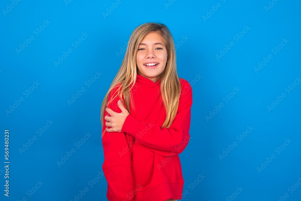 People, lifestyle, youth and happiness concept. Shy pretty caucasian teen girl wearing red sweater over blue studio background , feeling happy hugging herself. People, lifestyle, youth and happiness concept. Shy pretty caucasian teen girl wearing red sweater over blue studio background , feeling happy hugging herself.