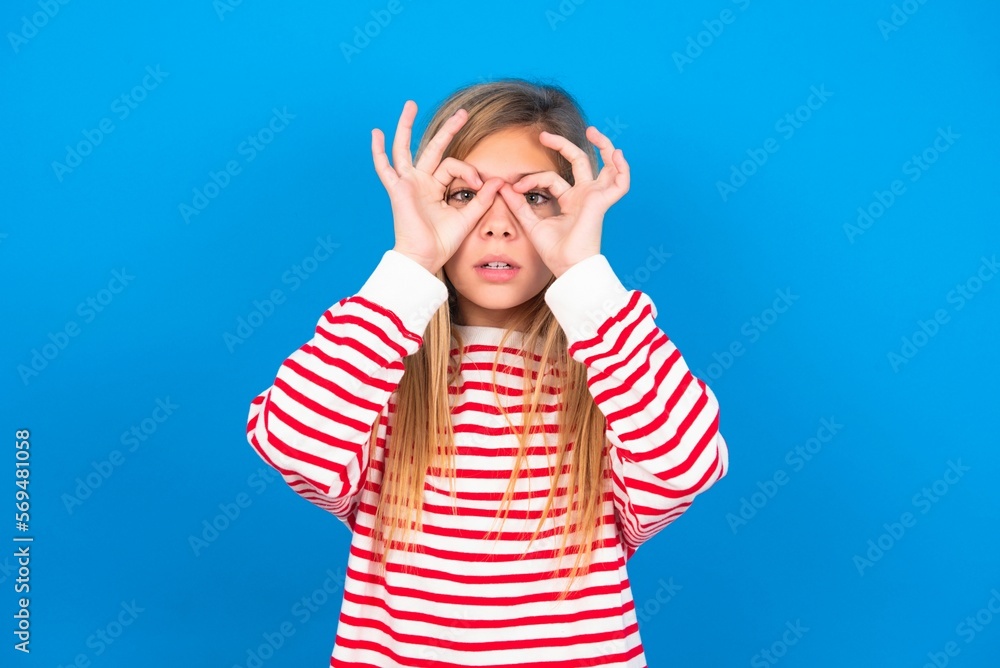 Playful excited caucasian teen girl wearing striped shirt over blue studio background showing Ok sign with both hands on eyes, pretending to wear spectacles. Playful excited caucasian teen girl wearing striped shirt over blue studio background showing Ok sign with both hands on eyes, pretending to wear spectacles.