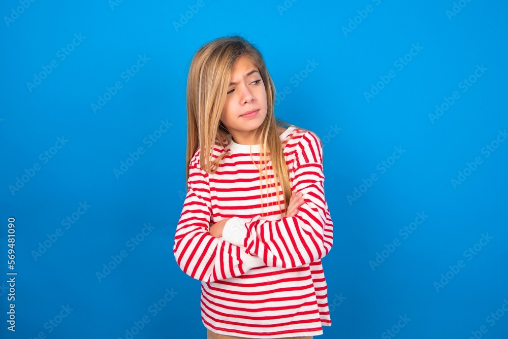 Displeased caucasian teen girl wearing striped shirt over blue studio background with bad attitude, arms crossed looking sideways. Negative human emotion facial expression feelings. Displeased caucasian teen girl wearing striped shirt over blue studio background with bad attitude, arms crossed looking sideways. Negative human emotion facial expression feelings.