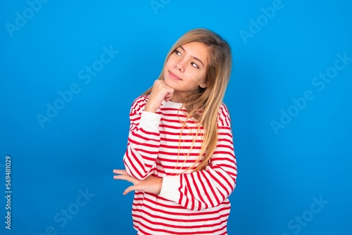 Face expressions and emotions. Thoughtful caucasian teen girl wearing striped shirt over blue studio background holding hand under his head, having doubtful look.