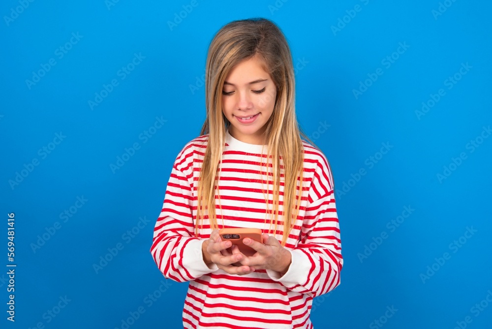 Smiling caucasian teen girl wearing striped shirt over blue studio wall using cell phone, messaging, being happy to text with friends, looking at smartphone. Modern technologies and communication. Smiling caucasian teen girl wearing striped shirt over blue studio wall using cell phone, messaging, being happy to text with friends, looking at smartphone. Modern technologies and communication.