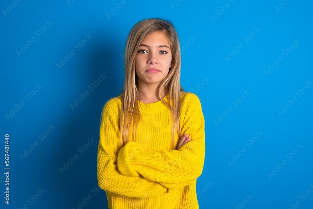 Self confident serious calm caucasian teen girl wearing yellow sweater over blue studio background stands with arms folded. Shows professional vibe stands in assertive pose. Self confident serious calm caucasian teen girl wearing yellow sweater over blue studio background stands with arms folded. Shows professional vibe stands in assertive pose.