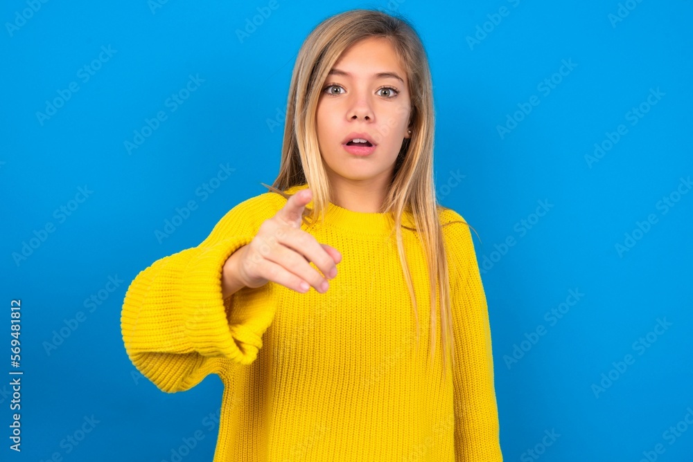 Shocked caucasian teen girl wearing yellow sweater over blue studio background points at you with stunned expression Shocked caucasian teen girl wearing yellow sweater over blue studio background points at you with stunned expression