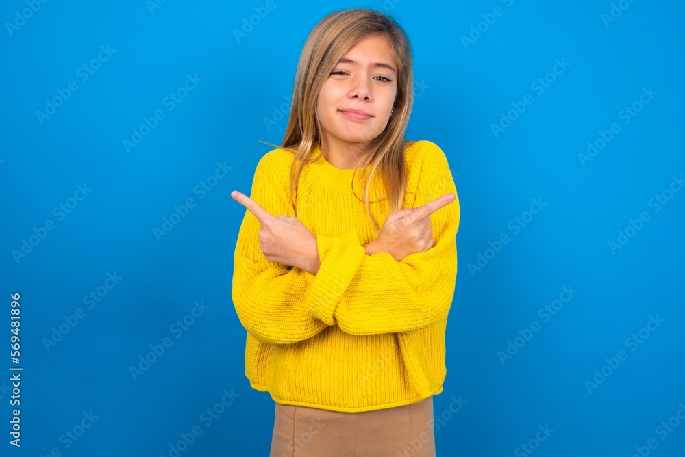 caucasian teen girl wearing yellow sweater over blue studio background crosses arms and points at different sides hesitates between two items or variants. Needs help with decision caucasian teen girl wearing yellow sweater over blue studio background crosses arms and points at different sides hesitates between two items or variants. Needs help with decision