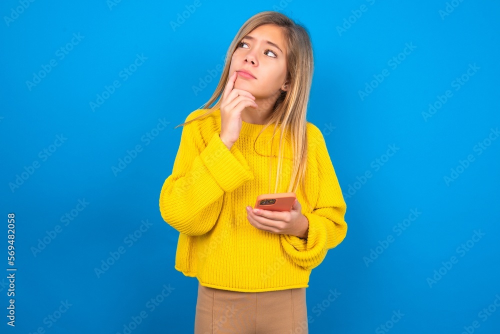 caucasian teen girl wearing yellow sweater over blue studio background thinks deeply about something, uses modern mobile phone, tries to made up good message, keeps index finger near lips. caucasian teen girl wearing yellow sweater over blue studio background thinks deeply about something, uses modern mobile phone, tries to made up good message, keeps index finger near lips.